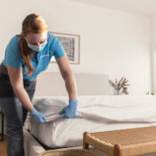 a maid setting up a white bedsheet of room, wearing a mask and gloves.